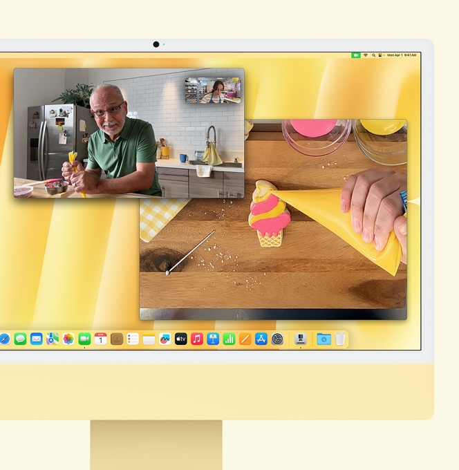 Dzeltens iMac. Desk View attēlojums. Ekrānā redzams maiznieks videozvanā ar citu personu. Vienā ekrānā redzami maiznieks un vīrieša seja. Otrā ekrānā redzamas maiznieka rokas, kas garnē cepumu.