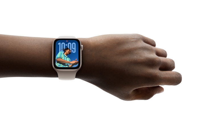 صورة مقرّبة لساعة Apple Watch باللون الفضي مع حزام مطاطي على معصم، تُظهر تطبيق الوقت بتصميم Liquid Glass الجديد