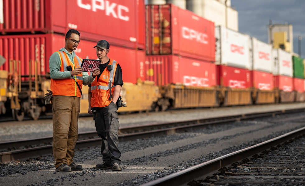 Deux cheminots dans une gare consultent un iPad à côté d’un train à l’arrêt transportant des conteneurs