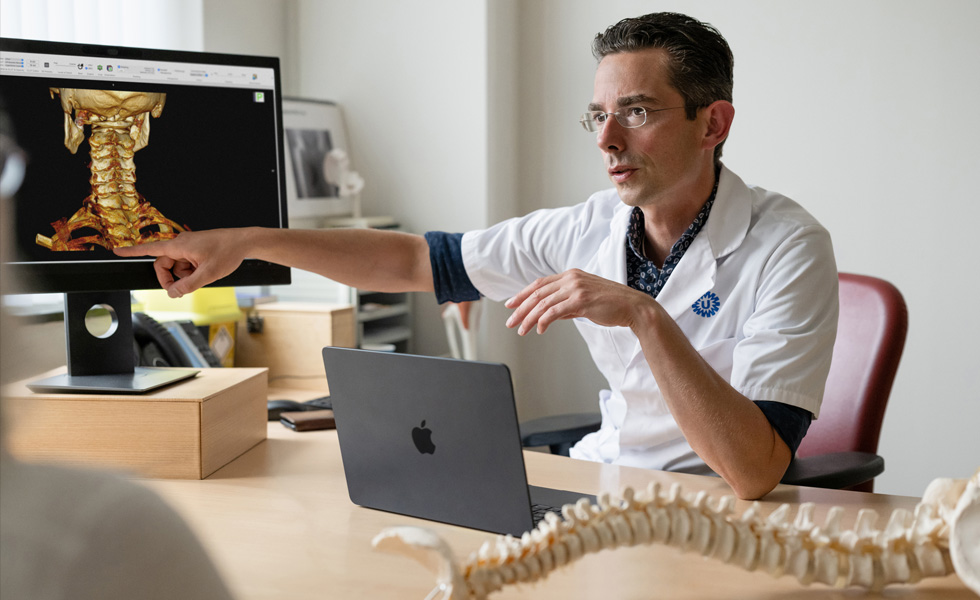 Ein Mediziner zeigt auf eine 3D-Skeletaldarstellung des Nackens auf einem Studio Display, während ein MacBook auf dem Schreibtisch steht